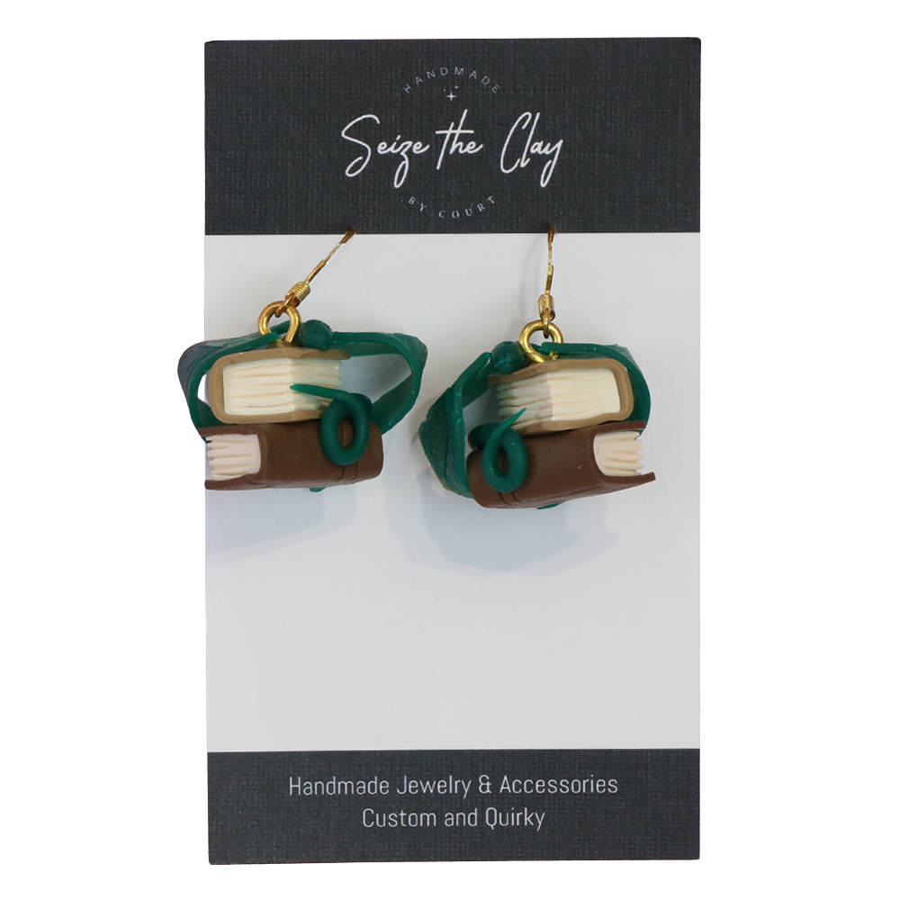 Book-shaped earrings with dragons wrapped around them, on a 'Seize the Clay' branded card