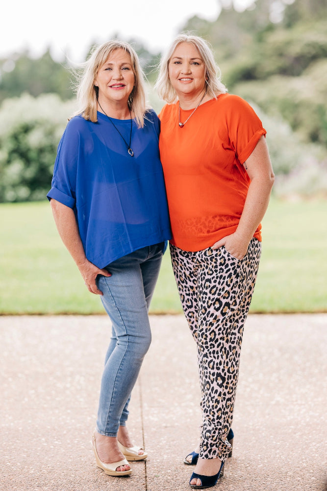 Two women standing outdoors, one in a blue top and jeans, the other in an orange top and leopard print pants.