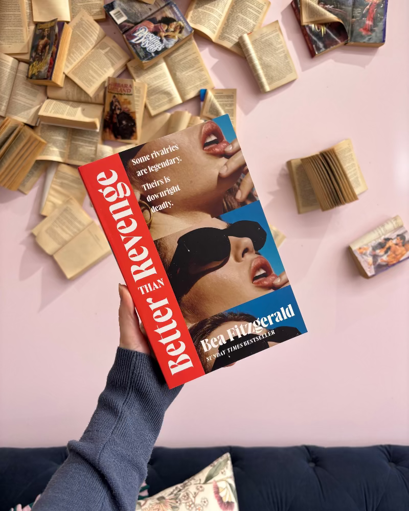 A hand holds a copy of the book 'Better than Revenge' by Bea Fitzgerald. The book cover features a red spine with white text, a blue section with the author's name, and a photo of a woman wearing black sunglasses.