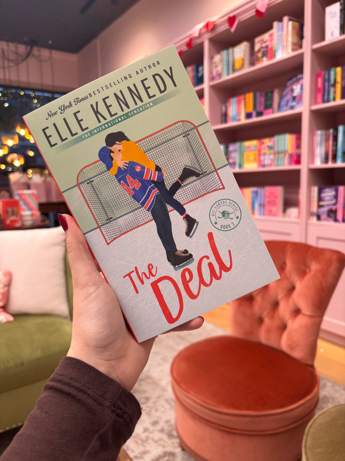Book titled 'The Deal' by Elle Kennedy held in a hand with a bookshelf background