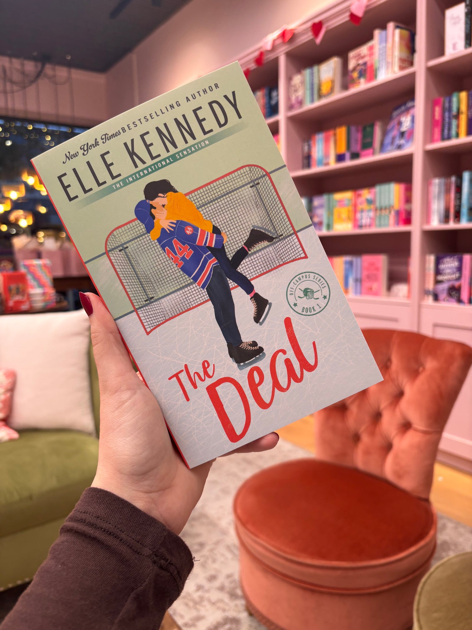 Book titled 'The Deal' by Elle Kennedy held in a hand with a bookshelf background